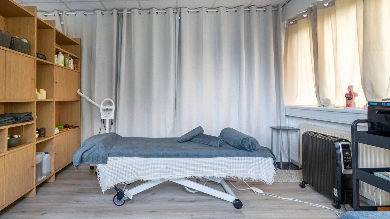 De behandelruimte: een opgemaakte massagetafel voor grijze gordijnen, eiken kast links, warm licht door het raam